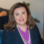 Kristy, a woman with golden brown shoulder-length hair, wears a purple blouse, navy blazer and white beaded necklace.