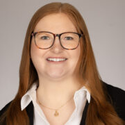Lilly, a white woman with straight auburn hair and brown glasses, wears a white blouse and black jacket.