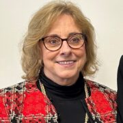 Nancy, a white woman with light brown hair and brown round glasses, wears a black turtleneck and red plaid blazer. 