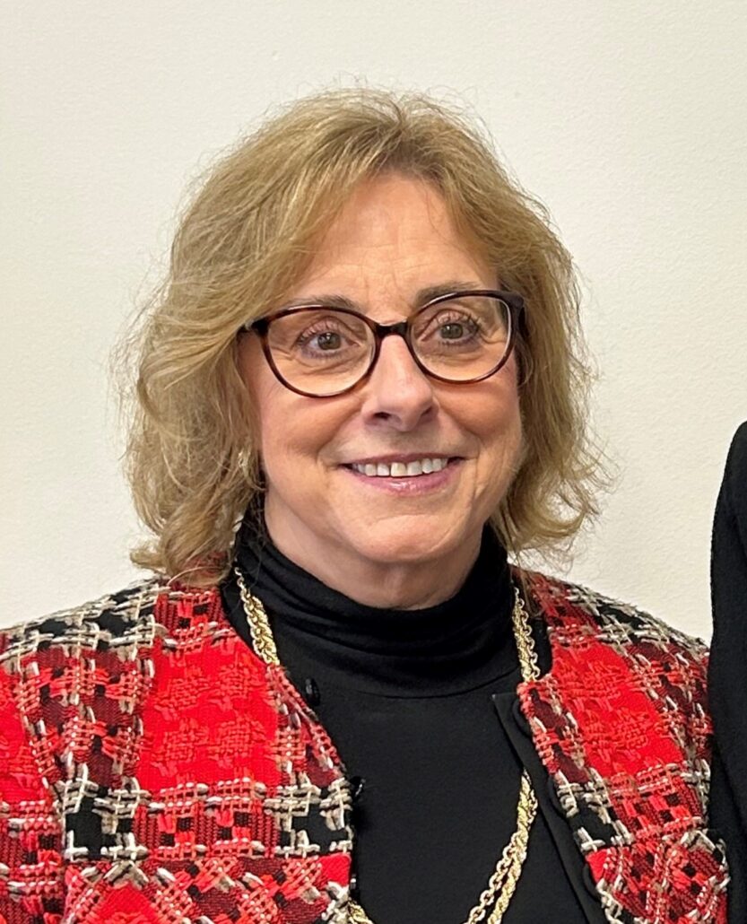 Nancy, a white woman with light brown hair and brown round glasses, wears a black turtleneck and red plaid blazer.