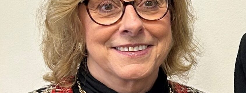 Nancy, a white woman with light brown hair and brown round glasses, wears a black turtleneck and red plaid blazer.