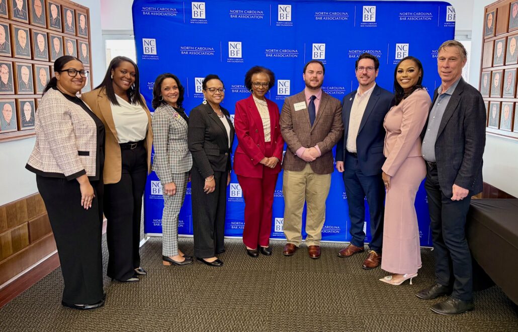 Alex is a Black woman with black hair. She is wearing a pink blazer and black pants. She is standing with the panelists, who are dressed in business attire and stand in front of a blue background with the NCBA logo on it. 