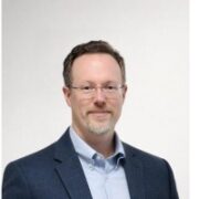 Sean, a white man with brown hair and clear glasses, wears a blue shirt and grey jacket. 