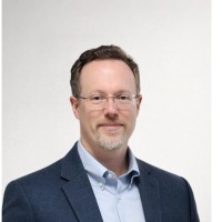Sean, a white man with brown hair and clear glasses, wears a blue shirt and grey jacket.