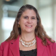 Susan, a white woman with brown hair, wears a black blouse and pink jacket. 