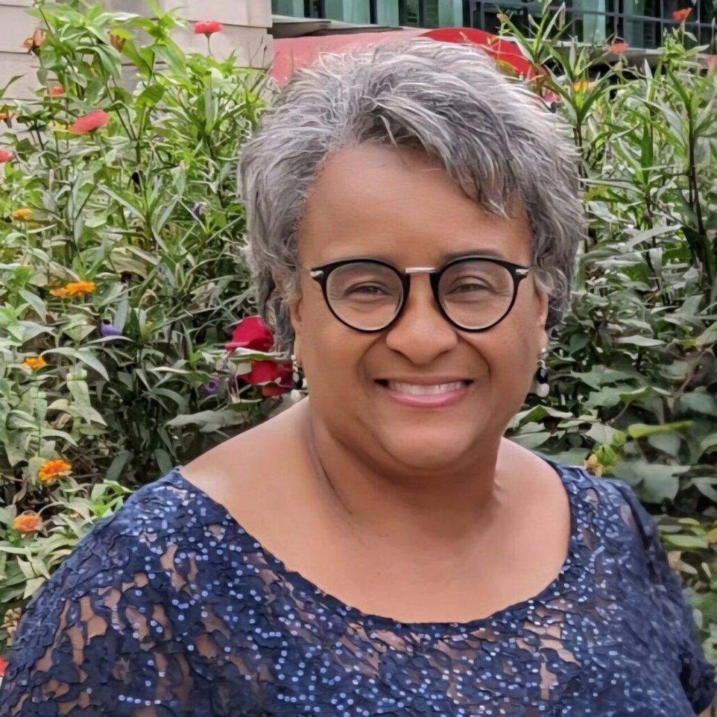 Bernice, a Black woman with grey hair, wears a blue dress with a floral lace pattern. She is standing outside with flowers behind her.
