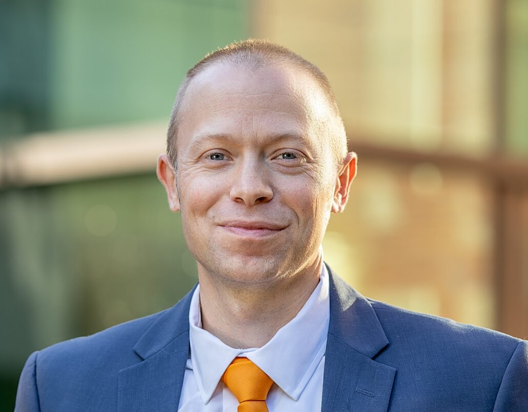 Matt, a man with light brown hair, wears a light blue shirt, orange tie and blue suit.