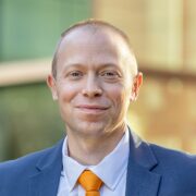 Matt, a man with light brown hair, wears a light blue shirt, orange tie and blue suit. 