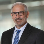 Jason, a white man with grey hair and black glasses, wears a white shirt, blue striped tie and black jacket. 