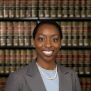 Chimara, a Black woman with black hair, wears a soft blue blouse, grey blazer and pearl necklace.
