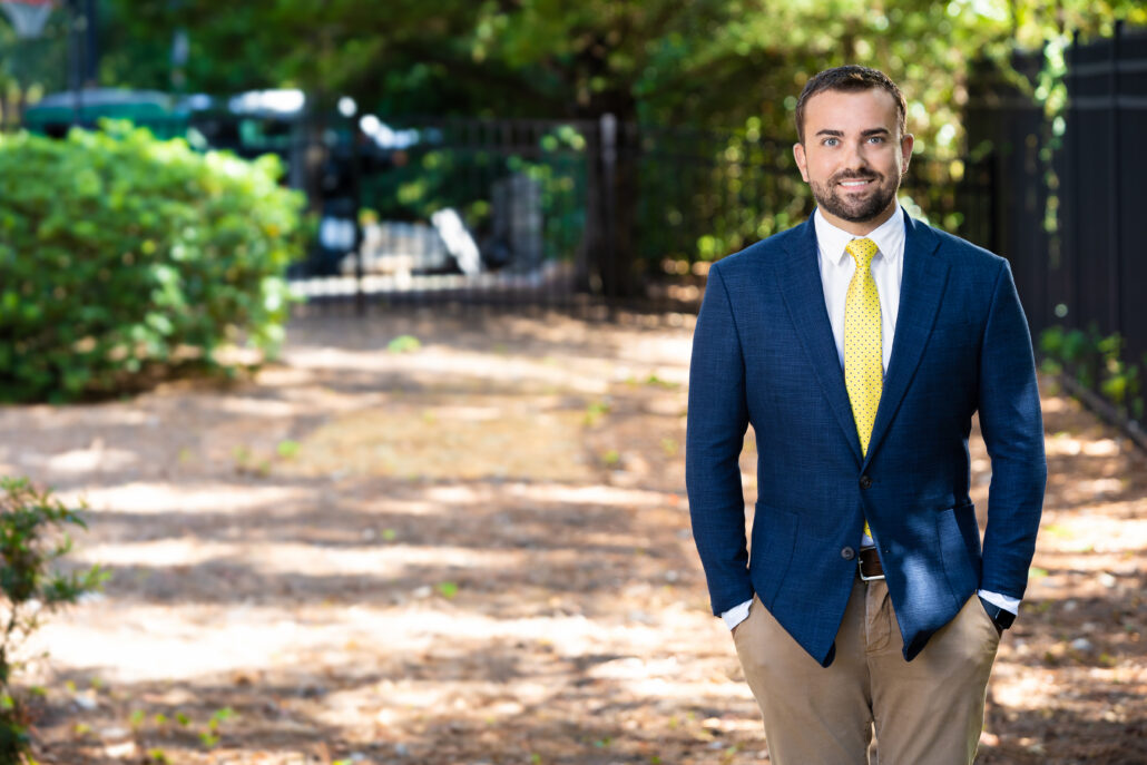 Cody, a white man with dark brown hair and a beard, wears a white shirt, yellow tie and blue suit.