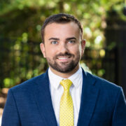 Cody, a white man with dark brown hair and a beard, wears a white shirt, yellow tie and blue suit.