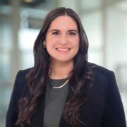 Ashley, a white woman with dark brown hair, wears a grey blouse and black blazer and gold necklace.