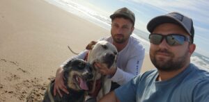 Cody, a man with brown hair and a beard, is wearing sunglasses. He is pictured with another white man with brown hair and a beard and they are with two dogs on the beach.
