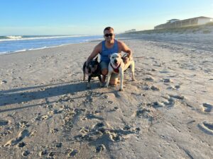 Cody, a white man with brown hair, is wearing a blue sleeveless shirt and is on the beach with his two dogs.