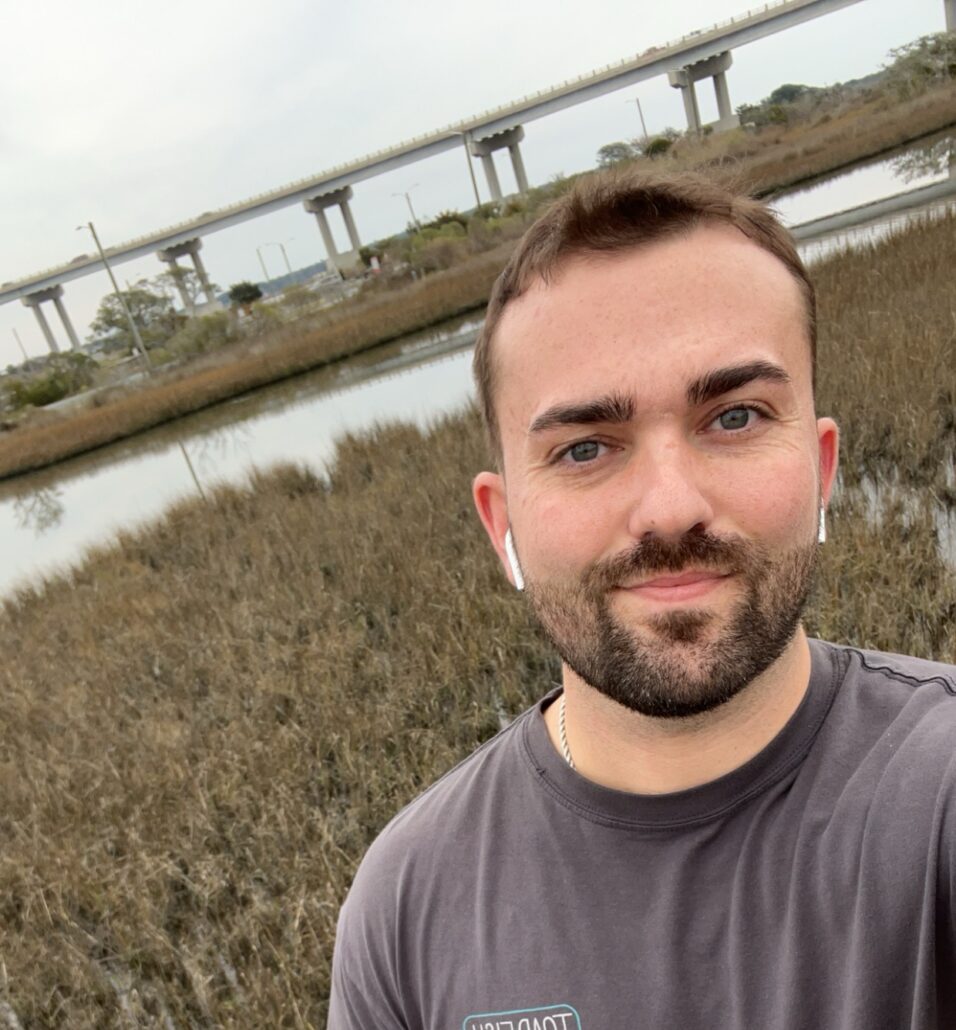 Cody, a white man with brown hair, wears a dark grey shirt and the beach is behind him.