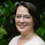 Maggie, a white woman with dark brown hair and glasses, wears an off white blouse.