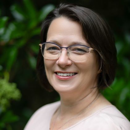 Maggie, a white woman with dark brown hair and glasses, wears an off white blouse.