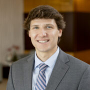 Harrison, a white man with brown hair, wears a pale blue shirt, blue and orange tie and light grey suit.