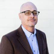 Robbie, a white man with a shaved head, wears black glasses, a blue and white checked shirt and a brown jacket.