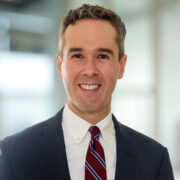 Joseph, a white man with brown hair, wears a white shirt, maroon tie with blue stripes and black jacket.