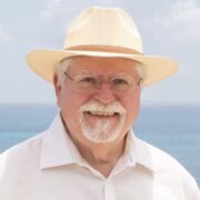 Douglas Wood, a white man with white hair and a beard, wears a white button down shirt, an ivory hat and clear glasses. The beach is behind him. 