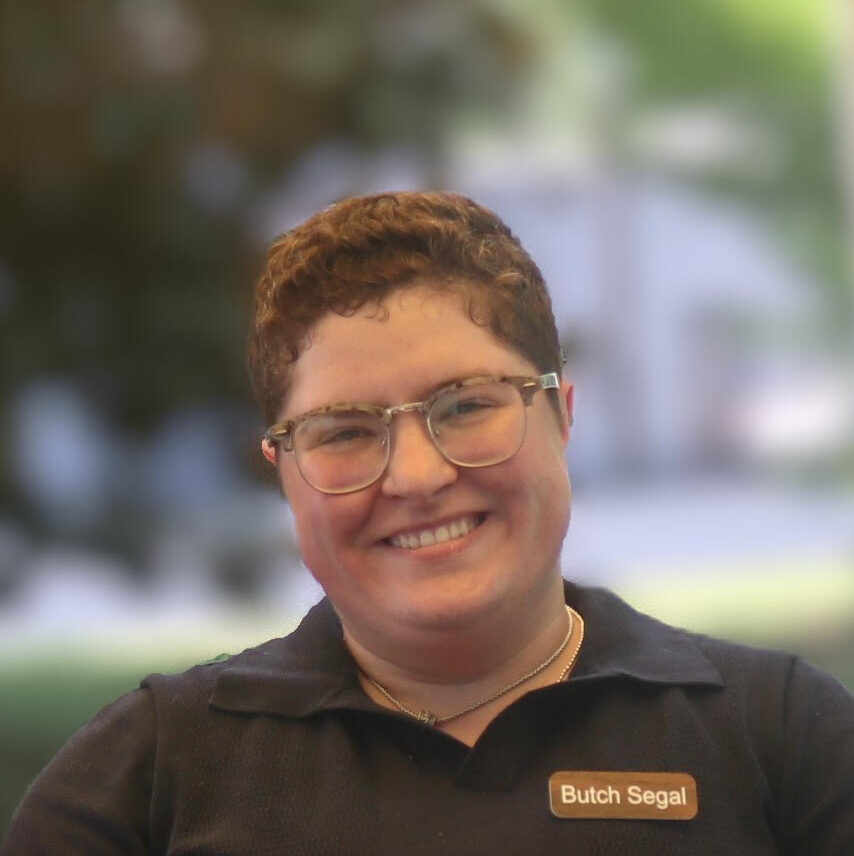 Butch, a white person with short brown hair, wears a dark brown polo with a nametag that that says "Butch Segal."