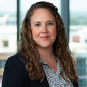 Chelsea, a white woman with wavy brown hair, wears a blue and white striped button-down blouse and a black suit.