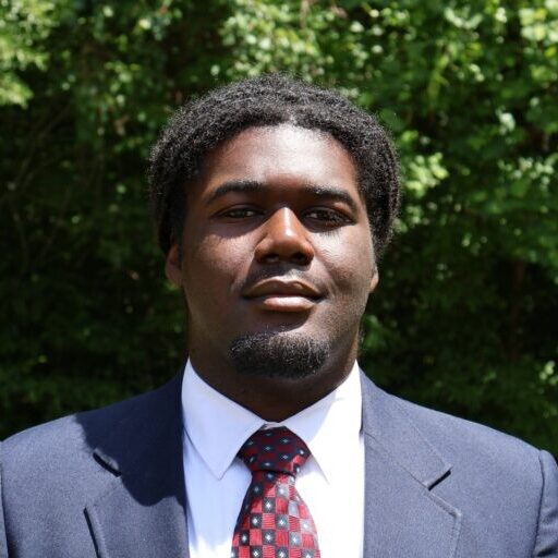 Richard, a Black man with Black hair, wears a white shirt, red tie and black suit.