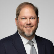 Robert, a white man with brown hair and a beard, wears a white shirt, grey tie and black suit. 