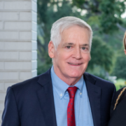 Gray, a white man with grey hair, wears a blue shirt, red tie and dark blue suit.