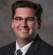 Karl, a white man with brown hair and glasses, wears a white shirt, dark tie and black suit.