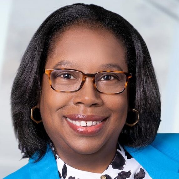 Tammy Nicholson is a Black woman with shoulder-length black hair and brown eyes. She is wearing a black and white patterned shirt, a bright blue blazer, and tortoise shell glasses. She is pictured smiling and standing against a white background.