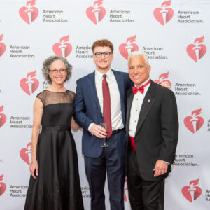 David wears a tuxedo with a white shirt and red bowtie, his spouse Cynthia wears a black dress, and his son wears a dark blue suit, white shirt and a red tie.