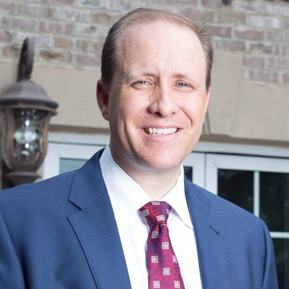 Adam a white man with light brown hair, wears a white shirt, red tie with light polka dots, and a white shirt. He is smiling.