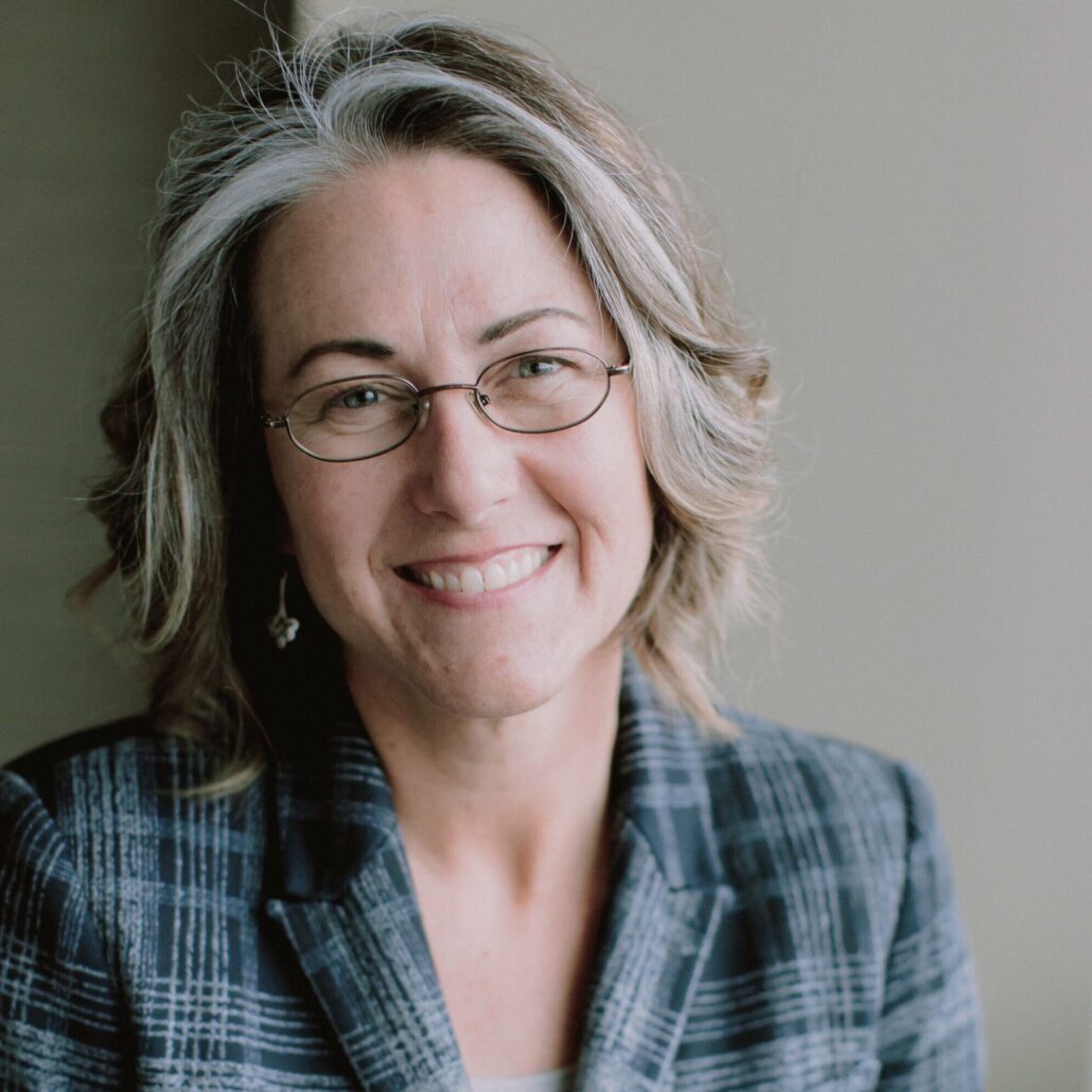 Amy, a white soman with short lond hair and glasses, wears a pale grey shirt and plaid grey blazer.