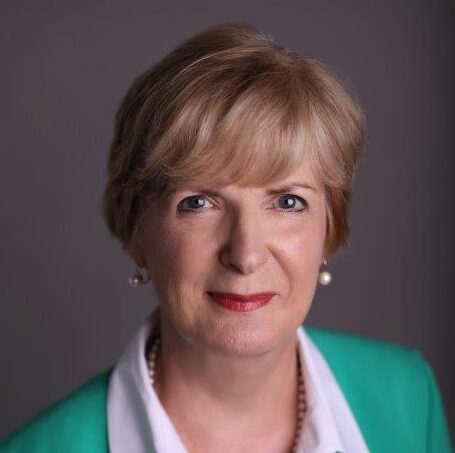 Ann, a white woman with short blond hair, wears a white blouse and teal jacket.