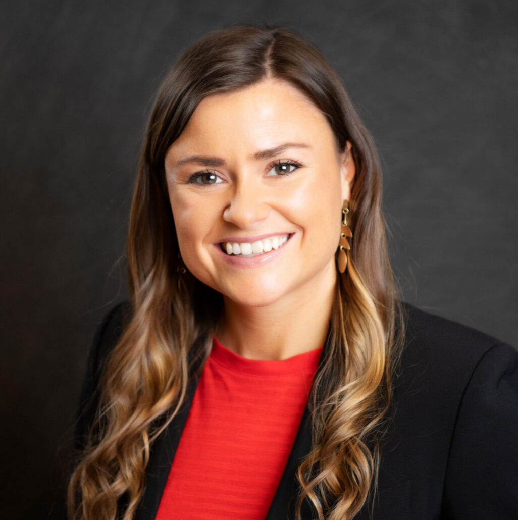 Ashley Banks is a young woman with golden brown hair and brown eyes. She is pictured smiling against a black background, and she is wearing a red shirt and a black blazer.