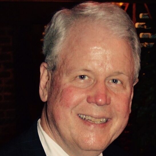 Bain, a white man with white hair, wears a white shirt and black suit.