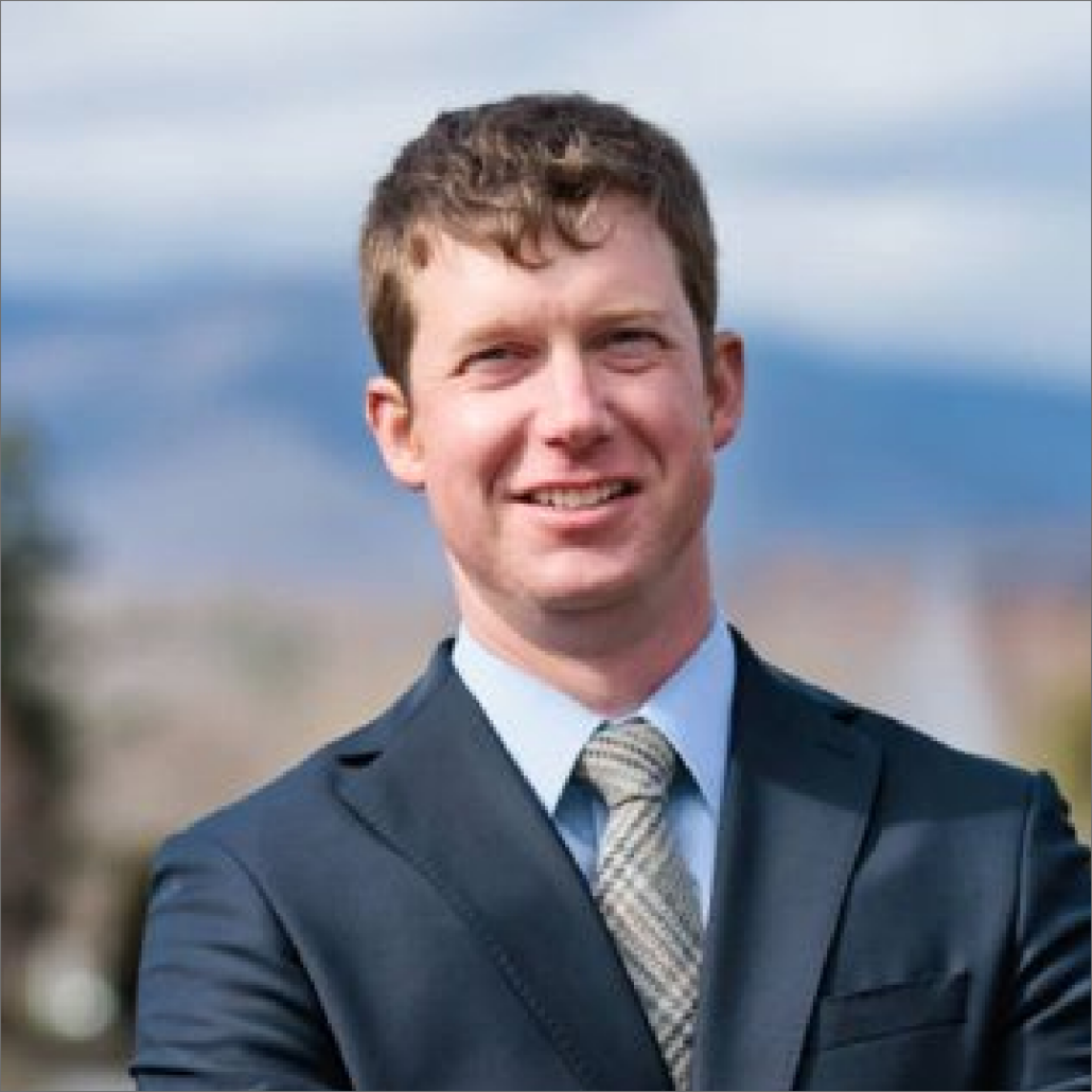 Barrett, a white man with brown hair, wears a blue shirt, grey plaid tie, and dark grey suit.