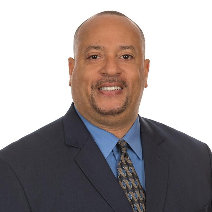 Brian Beverly is a Black man with a shaved head and a short brown beard and mustache. He has brown eyes. He is wearing a blue shirt and black tie with a white pattern and a black jacket.