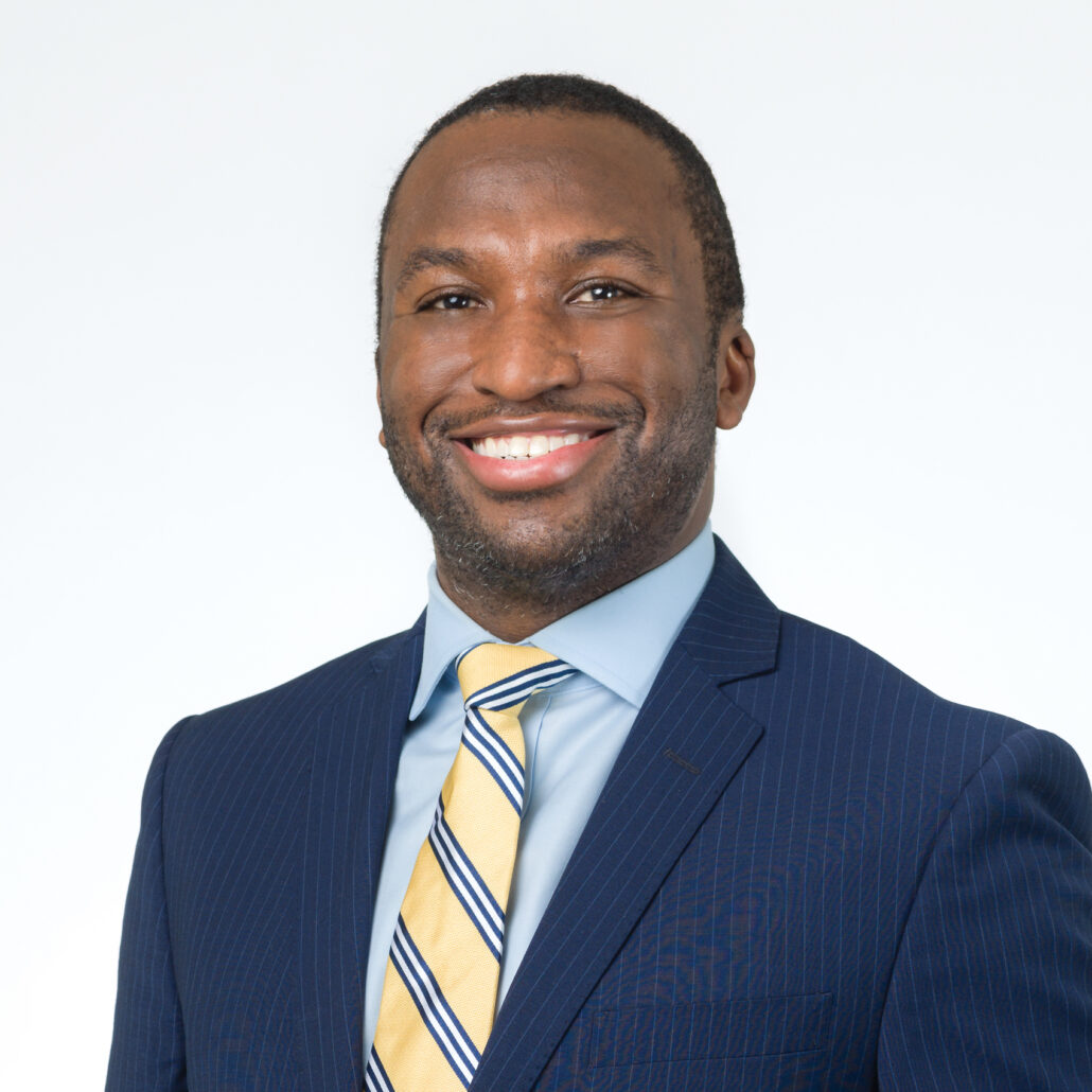 Jonathan, a Black man with black hair and a beard, wears a pale blue shirt, yellow and blue striped tie, and navy suit.