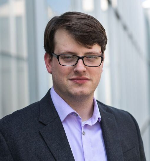 Brandon Huffman is a white man with brown hair. He is wearing black sqaure glasses, a pale blue shirt, and a black jacket. He is standing in the sun with a grey building behind him.