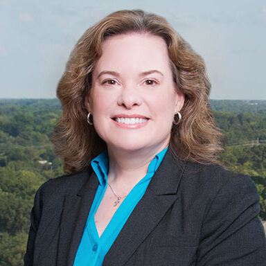 Christina Pearsall is a woman with wavy light brown, shoulder-length hair. She is wearing a bright blue shirt and a black blazer. She is standing outside and in front of green trees and a blue sky.