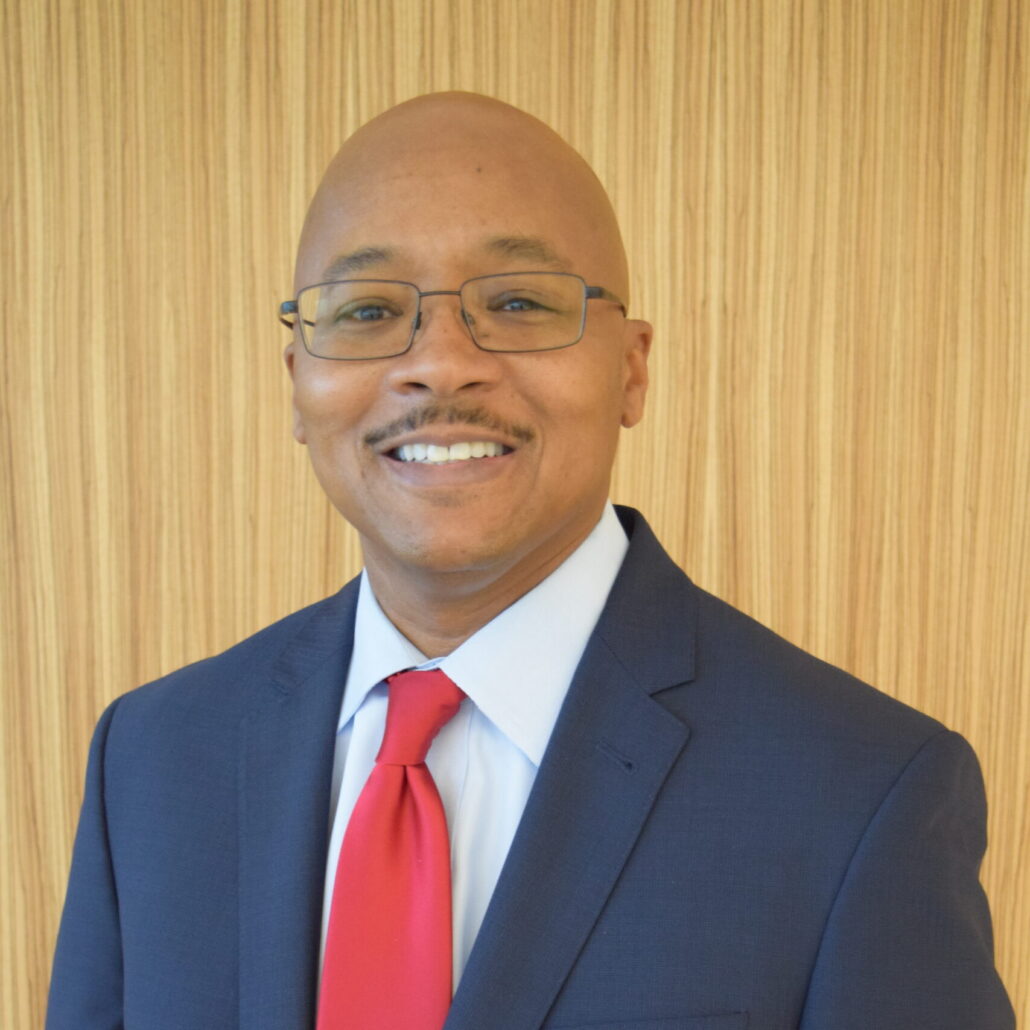 Clayton, a Black man, wears a white shirt, red tie and navy suit. He is smiling and standing with a wood-paneled wall behind him.