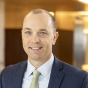 Paul, a white man with brown hair, wears a white shirt, green tie and dark grey suit.