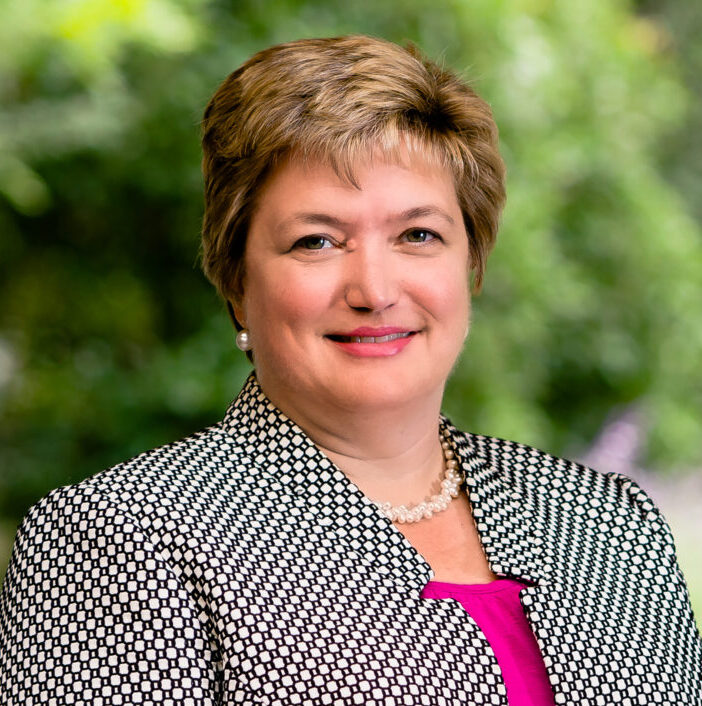 Crosby, a white woman with short golden brown hair, wears a pink blouse and black blazer with white dots.