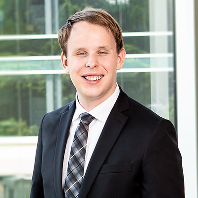 Derek, a white man with blond hair, stands before a sunny window and wears a black suit and gray plaid tie over a white shirt.