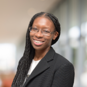 Cynthia, a Black woman with long black hair and glasses, wears a white blouse and dark grey suit.