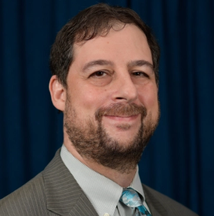 Eric, a white man with brown hair and a beard, wears a white sihrt, blue tie, and grey jacket.